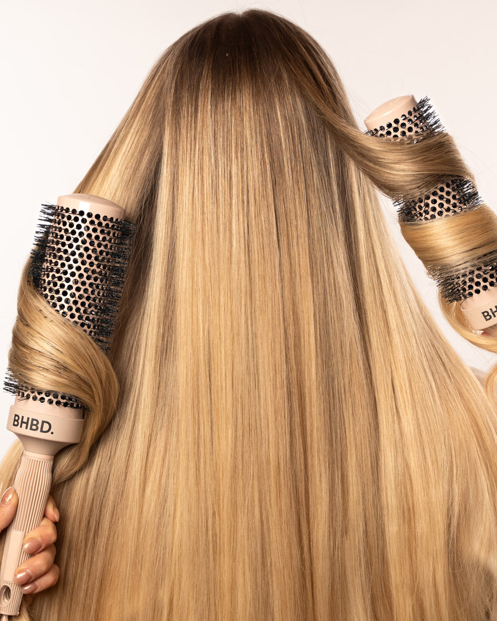 A person with long, straight blonde hair being styled with two BHBD round hairbrushes, showcasing a smooth and voluminous hair finish.